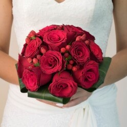 This detailed bridal close-up captures the moment a bride, seen from the waist up, pauses with her bouquet held carefully in front of her, as if waiting by the doors of a Stanmore venue before walking down the aisle. The bouquet itself is a meticulously crafted, rounded cluster of deep red roses, arranged so closely together that their velvety heads form a lush, almost cushion-like dome. Each rose varies slightly in openness: some buds are still tight and promising, while others have unfurled into full, spiralling blooms that draw the eye into their dark, romantic centres. Between these roses, small groups of vivid red hypericum berries peek through, their smooth, rounded surfaces offering a gentle contrast in shape and a fresher, lighter red tone layered into the design. The stems are hidden within a smart collar of broad, dark green leaves, folded and wrapped to create a polished, contemporary finish that still feels classic. The bride's hands, resting just below the flowers, hold the bouquet with a relaxed, reverent grip, suggesting both excitement and composure. Her white wedding dress, likely sourced from a local Stanmore or nearby Harrow boutique, is detailed with lace across the bodice and flows away in soft fabric, catching the diffused daylight. The background is softly blurred and neutral in tone, allowing the rich crimson and deep green of the bouquet to command attention. This composition speaks strongly of romance, passion, and commitment, a traditional red rose bridal bouquet that would feel perfectly at home in any Stanmore ceremony, from intimate gatherings near Stanmore Hill to grand celebrations closer to Canons Park.