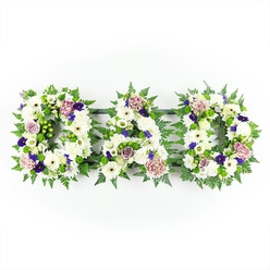 In this image, three substantial floral letters spell out DAD, arranged in a straight horizontal line and photographed against a perfectly plain white background, which allows every flower and leaf to be clearly seen. The letters themselves are constructed from a dense blanket of white chrysanthemums, their compact petals forming a textured but even surface that feels soft on the eye, like a comforting embrace. Interspersed among the chrysanthemums are crisp white gerbera daisies, their open faces and distinct centres adding gentle points of focus, while white lisianthus and small spray roses contribute elegant, layered blooms that break up the uniformity just enough to create subtle interest. Encircling each letter, a generous edging of deep green foliage gives the tribute structure and definition. Fine fern fronds and other leafy greens frame the white flowers and push them forward visually, while small clusters of bright green button chrysanthemums or berry-like accents tuck into the greenery to add contrast and a sense of freshness. Throughout the arrangement, carefully spaced touches of colour appear: deep violet and purple blossoms, similar to pansies or spray chrysanthemums, introduce a dignified richness, and softer shades of lavender and mauve carnations or roses weave in a note of gentle affection. The light is even and bright, with no harsh shadows, evoking the calm clarity of a still morning in Stanmore, perhaps before a service at a nearby church or cemetery. This DAD floral lettering feels both traditional and personal, a meaningful choice for families in the Stanmore area seeking a quiet but expressive way to say goodbye.