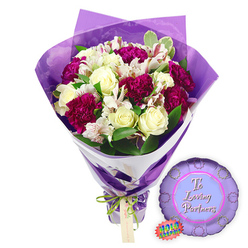 Shown against a simple white backdrop, this image captures a thoughtful graduation gift set, as if prepared by a Stanmore florist ready to be taken to a celebration meal on The Broadway. The main focus is a hand-tied bouquet wrapped in rich, velvety purple paper. The wrapping is folded with precision into elegant, sweeping panels that rise behind the blooms, then pulled in tightly at the base. A decorative green and white ribbon, which appears slightly patterned, is tied around the stems in a secure bow, creating a polished, presentation-ready finish. The flowers emerging from this wrapping form a full, rounded arrangement. Creamy white roses sit proudly among the other blooms, their petals smooth and tightly layered, suggesting freshness and formality. Nestled beside them are generous clusters of deep fuchsia or magenta flowers, likely carnations or chrysanthemums, whose numerous ruffled petals overlap to form plush, cushiony heads that bring warmth and energy to the design. Sprinkled throughout are fine stems of tiny white filler flowers that break up the bolder tones, giving the arrangement a soft, cloud-like feel. Surrounding foliage in varying greens lifts the entire palette, echoing the leafy avenues and gardens around Stanmore. Just below and to the right of the bouquet lies a round purple Mylar balloon. Its metallic surface reflects subtle highlights, and across the front, in graceful white script, are the words "To a Loving Graduate," making it unmistakably suited to academic celebrations. A small oval logo or tag, trimmed in green and white, appears next to the balloon, suggesting the mark of a trusted local florist. Together, bouquet and balloon convey admiration, pride, and encouragement-perfect for congratulating a new graduate returning home to Stanmore after their big day.