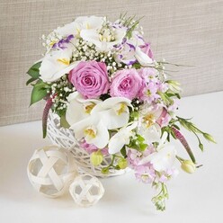 A richly detailed wedding-style centerpiece in shades of pink and white fills the frame, designed as if for an intimate celebration in Stanmore. The flowers are arranged in a round white wicker basket that sits on a gleaming white surface, immediately drawing the eye to the interplay of textures and colours. Central to the design are several lush pink roses, each bloom in full, generous opening, with petals curling in graceful spirals from the tightly packed heart out to the softer, more relaxed edges. Their gentle blush tones radiate romance and create a focal point that anchors the whole composition. Draped around and among them, white Phalaenopsis orchids extend their wide, porcelain-like petals, the centers glowing with warm yellow and soft apricot hues touched with a hint of lilac, evoking the refined atmosphere of a reception at a Stanmore country club or stylish restaurant. White Lisianthus blossoms, some edged in pale lavender, add a fluttering, ruffled element, enhancing the piece's sense of depth and delicate movement. Throughout the arrangement, airy sprigs of white baby's breath create a fine, lacy network that softens transitions between blooms and adds a sense of lightness, as if the flowers are floating. Pale pink stock flowers rise on slender stems, their clustered florets adding both height and a gentle cascading line that falls over the basket rim, mirroring the natural drape of a bridal bouquet. Fresh green foliage, peeking between petals and trailing slightly at the edges, introduces a vivid counterpoint to the pastels, suggesting newly cut stems and garden freshness. On the tabletop beside the basket, two woven decorative spheres in pale natural fibre sit quietly, their twisted strands echoing the basket's craftsmanship. A neutral, light beige fabric backdrop frames the entire scene, allowing the details and colours of the flowers to stand out. The mood is one of elegant joy, ideal for weddings, anniversaries, or special gatherings in and around Stanmore.