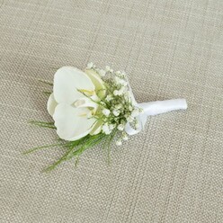 A refined white orchid boutonniere is carefully placed on a pale beige, woven fabric surface, whose subtle texture lends a rustic-luxe backdrop reminiscent of a well-appointed Stanmore townhouse. The eye is immediately drawn to the central Phalaenopsis orchid, its petals broad and satin-smooth, opening in a tranquil, symmetrical curve. The pure white of the bloom is kissed with a hint of buttery yellow deep in its throat, a small, sunlit heart that gives warmth and dimension to the otherwise cool tones. Around this central flower, an airy ring of baby's breath is arranged in delicate sprays, each stem tipped with clusters of miniature white blossoms. These tiny gypsophila flowers form a misty halo, softening the silhouette of the orchid and creating a romantic, almost ethereal effect, as if the boutonniere carries its own gentle glow. Slender fronds of fine green foliage weave through and beneath the white flowers, their feathery lines adding movement and a fresh, garden-picked character that feels at home in the leafy streets near Stanmore Country Park or along Marsh Lane. The stems are drawn neatly together at the base and wrapped with a bright white satin ribbon, the binding smooth and precise, finished with a small, elegant bow that signals the piece is ready to be fastened securely to a jacket lapel. The lighting is soft and even, accentuating every detail without harshness, so that the textures of petal, ribbon, and fabric are all clearly defined yet harmonious. This boutonniere evokes a sense of understated luxury and timeless romance, perfectly suited to a groom walking into a Stanmore wedding ceremony, a prom date arriving in north London, or any formal occasion where a single, beautifully crafted flower can quietly express grace, devotion, and thoughtful preparation.