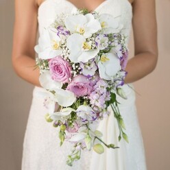 This poetic image from Stanmore tells of a bride cradling a magnificent cascading bouquet, set vividly against a tranquil beige background evocative of early morning light filtering across Stanmore Broadway. The bouquet, sculpted into a thoughtful teardrop, overflows with layered tones of pure white, tender pink, and mystical lavender. Majestic phalaenopsis orchids, their large pristine petals and sunny yellow centers catching soft light, are harmoniously paired with plush pink garden roses-each rose a swirl of velvety petals that speaks to timeless romance. Clusters of delicate lavender stock, their slender blossoms, and ruffled eustoma with purple-tipped edges provide textures as varied as the flowerbeds of Bentley Priory Nature Reserve. Baby's breath adds gentle clouds of whiteness throughout the bouquet, enhancing its fullness with a whisper-light touch, while discreet green foliage peeks through in quiet support. The bride's white lace bodice and gentle embrace focus all eyes on this exquisite composition-a floral celebration crafted by skilled local florists of Stanmore. A soft, clean fragrance and serene presence offer a fitting tribute to love, making this bouquet perfect for weddings in the historic and leafy heart of north-west London.