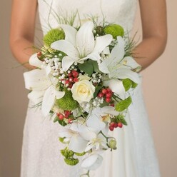 Cradled beneath the soft folds of an ivory lace gown, this exquisite bridal bouquet radiates the gentle luxury and timeless beauty that Stanmore couples so often desire. Prominent white lilies, their broad petals pearly and speckled with touches of russet, anchor the bouquet and create an air of classic sophistication. At the centre, a creamy white rose unfolds in tender whorls, surrounded by feathery baby's breath that seems to float, much like morning mist gathering along the trails of Bentley Priory Nature Reserve. Bold pops of lime-green cushion mums invigorate the composition, infusing the design with cheerful modernity reminiscent of the vibrant gardens found throughout Stanmore. Glossy red hypericum berries add a splash of playful colour and a festive spirit-recalling the bustling charm of Stanmore's local flower markets. Lively tendrils of fine greenery weave naturally among the blooms, their movement reflected in the bouquet's dramatic teardrop cascade of white Phalaenopsis orchids, lending both flow and effortless elegance. Every petal is illuminated by soft daylight, revealing subtle textures and a gentle harmony, the mark of an experienced Stanmore florist's handiwork. This arrangement is perfectly suited to romantic wedding ceremonies in Stanmore's historic venues, promising freshness and artistry for every special moment. Same-day delivery can be arranged across the area for those seeking a bouquet infused with local grace.