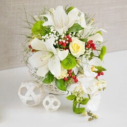 Captured in soft, even light, this floral arrangement exudes the quiet elegance often requested for Stanmore weddings and special occasions. The flowers are gathered in a low, round basket of white woven wicker, resting on a pristine white surface that enhances the freshness of the blooms. From the basket rises a generous composition dominated by large white lilies, their open trumpet flowers facing in different directions as if greeting guests around the table. The lily petals are smooth and luminous, with subtle speckles near the centre and pale stamens that suggest a gentle, powdery fragrance. Nestled just off the centre, a single cream-to-light-yellow rose forms a soft focal point, its closely packed petals unfolding in a neat spiral, adding a hint of warmth among the cool whites. Flowing from the right side of the arrangement, a delicate cascade of white phalaenopsis orchids drifts downward, each bloom with broad petals and a gracefully curved profile, echoing the movement of a traditional cascading bridal bouquet often seen at ceremonies in Stanmore's local venues. The bouquet is richly layered with foliage: fine, wispy fronds, broader green leaves, and bright green pom-pom flowers that resemble tiny cushions, providing intriguing texture and depth. Speckles of tiny white baby's breath appear between the larger blooms like a soft haze, while glossy ruby-red berries are tucked here and there, bringing small bursts of colour that feel celebratory and seasonal, suitable for a festive dinner near Stanmore Hill. The background is a neutral, textured beige surface, calm and understated, ensuring the flowers remain the star. To the left of the arrangement, two white decorative spheres made from interlaced strips sit on the tabletop, mirroring the basket's weave and adding a contemporary, sculptural touch. The composition feels refined, fresh, and thoughtfully composed, as if crafted by an experienced local Stanmore florist with a keen eye for balance and detail.