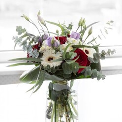 Soft natural light spills across an exquisite bouquet set against the serene backdrop of Stanmore, imparting a sense of tranquil elegance. Luscious red roses at the centre evoke passion and warmth, their deep colour juxtaposed by the crisp purity of white gerberas, each crowned with a rich, chocolate-brown eye. The arrangement has a gentle, contemporary allure, thanks to sprigs of light mauve lisianthus that stretch upward, some still shyly curled in bud. The bouquet's structure is elevated by airy sprays of delicate white filler, giving a sense of lightness and cloud-like volume. Framing the blooms, sprigs of eucalyptus add a silvery-green sheen and a distinct, refreshing scent, reminiscent of Stanmore's wide-open green spaces. The arrangement is bordered by broad, glossy foliage that wraps tightly around the base, providing both polish and support. From the transparent vase, fresh stems are visible, anchored in sparkling water-a testament to local freshness and florist care. Suited to mark new beginnings, deliver comfort, or celebrate cherished milestones, this bouquet brings a touch of Stanmore's refined grace and natural beauty into any home.