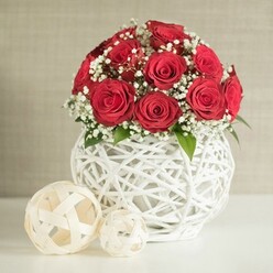 This photograph captures a sophisticated floral centrepiece built around a compact dome of red roses, their petals unfolding in dense, velvety spirals that speak of passion and timeless elegance. The blooms are tightly grouped, forming a smooth, rounded silhouette that draws the eye to the interplay of light and shade within each flower. Interwoven throughout the roses are threads of white baby's breath, their tiny clustered blossoms creating a soft, mist-like veil that lightens the arrangement and adds a sense of movement. Occasional glimpses of fresh green foliage peek through the flowers, grounding the design with natural contrast and suggesting careful, expert conditioning. The bouquet is cradled in a rounded white vase formed from interlacing strands that twist and loop around one another in an open-weave structure, much like a contemporary wicker or rattan sculpture. This unique vessel, bright and clean, provides both texture and modern flair, making it as much a part of the visual story as the flowers themselves. To the left on the smooth white surface are two decorative orbs fashioned from light, cream-coloured strips woven into airy lattices, the larger sphere positioned slightly forward to create depth. A neutral, fabric-like wall in soft beige forms the background, blurred just enough to keep attention on the central arrangement. The scene feels like a carefully curated corner of a Stanmore dining room or wedding venue near Stanmore Hill, ideal for anniversaries, intimate celebrations, or romantic gestures, with each element suggesting the steady hand of an experienced florist at Flowers Stanmore.