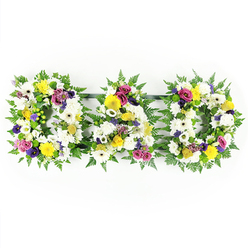 This image shows a poignant "DAD" floral tribute, thoughtfully arranged for a father's funeral or memorial service and set against a bright white backdrop so that every flower is clearly defined. Three bold letters - D, A, and D - are aligned horizontally on thin dark green supports, visually linked yet each complete in its own right. Around the edge of each letter, lush green fern leaves fan outward, their soft, feathery texture adding a natural outline reminiscent of the greenery around Stanmore Hill and local gardens. The central area of the letters is tightly packed with fresh white blooms: rounded chrysanthemums and large white gerbera daisies form a serene base, their layered petals creating a cushion-like effect. Nestled amongst these are golden-yellow gerbera heads and delicate yellow spray roses, bringing gentle warmth and optimism that softens the formality of the white. Rich purple lisianthus blossoms and clusters of magenta or deep pink spray roses appear at intervals, their deeper tones suggesting affection, admiration, and cherished memories. Tiny lavender flowers and crisp green button chrysanthemums peek through in places, adding fine detail and lively contrast. The arrangement is clearly the work of an experienced florist, with each bloom carefully angled to face forward, ensuring the word "DAD" reads clearly from a distance in a church, crematorium, or graveside in and around Stanmore. The overall effect is of a respectful, carefully balanced sympathy tribute that honours a beloved father's life, combining calm whites, hopeful yellows, and heartfelt purples in a single, lovingly crafted design.