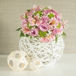 As I thoughtfully arrange these blooms in Stanmore, I can almost feel their varied textures: the classic velvet of each pastel pink and lavender rose, the crisp, smooth coolness of the white open-weave vase under my hands, and the gentle rustle of deep green leaves. The design-perfect for a wedding on Stanmore Hill or a milestone celebration near Whitchurch Gardens-blends artistry and comfort: petite pink alstroemeria with subtle striped faces nestle among rosy blooms, their soft trumpet shapes offering whispered elegance. Invigorating lime green button chrysanthemums dot the bouquet with playful pops of colour, while clouds of baby's breath fill the space with an airy, ethereal touch. The arrangement is lovingly presented in a white, rounded basket-its intricate woven pattern a nod to Stanmore's appreciation for handcrafted beauty and timeless design. To the left, two pale decorative spheres enhance the scene, their shapes and textures reminiscent of the gentle, rolling greens beyond Stanmore Cricket Club. All rest on a spotless white surface, backed by a softly textured, neutral-toned wall that draws out the bouquet's colour story. The overall mood is one of serene confidence and quiet joy, a centerpiece radiating both grace and heartfelt intention-inviting smiles and celebration in any Stanmore space.