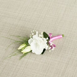 A refined floral boutonnière lies diagonally across a textured oatmeal-beige fabric, its subtle woven pattern reminiscent of natural linen and the understated elegance of Stanmore's quiet residential streets. At the centre of the composition is a fully opened white lisianthus bloom, its many fine, ruffled petals layered one upon another like delicate tissue, forming a soft, romantic cup of pure white. Nestled close to it, two slender lisianthus buds in creamy green tones tilt gently to the side, still closed yet clearly moments away from opening, a tender symbol of beginnings that feels especially apt for a wedding or engagement celebration. Feathery clusters of white gypsophila peek out around the main flower and buds, their tiny blossoms scattered like a dusting of snowflakes, adding airy volume and a sense of lightness. Behind and around the flowers, slim, grass-like fronds of greenery arch outward, stretching toward the left side of the frame, while a broader, velvety dark green leaf provides a confident backdrop that helps the white petals stand out. The stems of this small floral piece are neatly bound near the bottom with a vivid fuchsia pink satin ribbon, wrapped tightly and finished in a playful bow that introduces a bright, modern pop of colour. The satin catches the soft, diffused light, giving a slight sheen that contrasts beautifully with the matte fabric beneath. This carefully balanced boutonnière, ideal for a groom's lapel at a Stanmore ceremony or a formal event near Canons Park, radiates freshness, poise, and a gentle joy, as if capturing one perfect, hopeful moment in flowers.