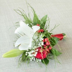 Set against a creamy, softly woven fabric reminiscent of the welcoming textures found in Stanmore's family homes, this vibrant floral arrangement tells a story of grace and celebration. The centrepiece is a luminous white lily, the petals open in full, kissed with faint russet speckles and glowing yellow anthers. Nearby, a plush red rose unveils its velvety spirals, symbolising enduring love and local elegance-evocative of the cherished memories made in the parks and venues around Stanmore. Scattered throughout, glossy red berries introduce bold, festive highlights, their sheen reminiscent of the spirited markets on The Broadway. Baby's breath airy clusters impart a gentle softness, enchanting the senses, while a slender green lily bud arcs out to the side, a suggestion of renewal and growth. The entire composition is wrapped in varying greens-delicate ferns and deep-leaved foliage lending a vibrant foundation. Brought together at the base with a luxuriant red satin ribbon, the bouquet is both artful and practical, suited perfectly to bridal gatherings and commemorative occasions throughout Stanmore. This arrangement, aglow in soft, ambient light, radiates freshness and heartfelt intent, ready for prompt delivery to mark life's most special celebrations.