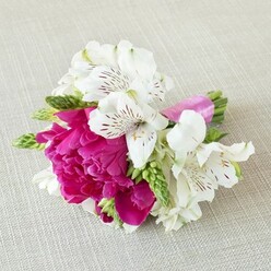On a pale beige, linen-like fabric that recalls the calm, understated interiors of Stanmore's family homes, a small yet luxurious floral arrangement rests horizontally, carefully composed but with an effortless grace. At its centre is a rich fuchsia bloom-most likely a compact peony or garden rose-its petals densely layered and softly ruffled, forming a rounded, plush head of colour that feels almost touchable. Surrounding this main flower are several white alstroemeria, or Peruvian lilies, each with six slender petals that sweep outward in a gentle arc. The inner petals are finely marked with maroon and deep red streaks and speckles, as if nature had written delicate calligraphy across their satiny surfaces. Threaded through the design are slim, elongated green buds and foliage, providing zesty pops of fresh green and a slightly spiky texture that contrasts beautifully with the softness of the main bloom. The stems of the flowers are gathered tightly and bound with a smooth, bright pink satin ribbon, its sheen catching the light and echoing the central fuchsia tones with stylish precision. The light in the image is soft and even, without harsh shadows, allowing every nuance of colour and every tiny vein in the petals to be appreciated. This compact piece feels perfect for a groom's boutonnière, a prom corsage, or a small hand-held posy, something you might choose for an intimate ceremony at a local Stanmore venue or a celebration near Queensbury. It evokes modern romance, joyful connection, and that special sense of occasion that local florists near Flowers Stanmore understand so well when creating bespoke, fresh arrangements for same-day delivery across the area.