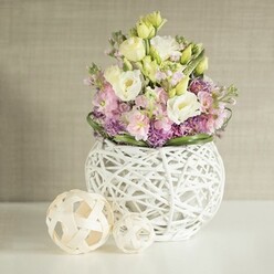 Bathed in serene daylight near Stanmore's Bentley Priory, a luxurious bouquet takes pride of place on a crisp white tabletop, its blooms artfully arranged to embody sophistication and natural allure. Creamy white and pale yellow eustoma, famed for their sumptuous, ruffled petals, are seamlessly paired with clusters of soft pink stock, whose verticality and gentle floral scent conjure memories of early walks through Stanmore Country Park. Lush purple filler blooms-possibly allium or asters sourced from nearby gardens-provide rich contrast beneath the pastel mainstays, anchoring the composition and creating a tactile patchwork that draws the eye. The entire arrangement is beautifully enclosed by a neat collar of fresh green leaves, heightening the sense of fullness and artisanal quality. At the base sits a striking white vase, its spherical form and intricate openwork rattan design communicating both modernity and the understated grace seen in Stanmore's interior style. Two decorative spheres, expertly woven from pale wood or rattan and positioned just to the left, echo the vase's organic patterns and tie the arrangement into a cohesive tableau. Behind it all lies a subtle backdrop-a warm, greige wall with delicate vertical fabric texture-serving as the perfect stage to let the colours sing. Florists from Flowers Stanmore have poured their expertise into this display, ensuring that each element harmonises to evoke themes of romance, celebration, and the enduring elegance found throughout Stanmore's welcoming neighbourhoods.
