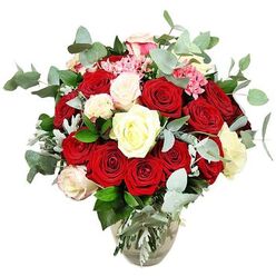 A lavish rose bouquet rises from a simple clear glass vase, photographed against a spotless white backdrop so every bloom appears crisp and defined. The composition features a generous collection of deep red and bright scarlet roses, their velvety petals layered and plush, some still cupped in bud and others wide open in full, romantic splendour. These rich tones are softened by an array of creamy white and ivory roses threaded throughout the arrangement, their paler shades reflecting the light and adding a sense of serenity and balance. A few of the white blooms are gently brushed with pale pink around the petal edges, introducing a shy hint of colour that bridges the gap between the vivid reds and the calm whites. Nestled among the roses, small clusters of bright pink filler flowers, resembling tiny carnations or miniature blossoms, contribute intricate texture and bring an uplifting playfulness to the bouquet. Encircling and supporting the roses, you see lush sprays of blue-green eucalyptus leaves, rounded and slightly silvery, offering a fresh, modern contrast and hinting at a clean, aromatic scent. Dark green foliage and subtle silvery-green leaves fill out the base and sides, ensuring the bouquet forms a complete, rounded dome with no empty patches. The glass vase itself flares at the base and narrows at the neck, with the stems neatly gathered and the water level clearly visible, giving the impression of freshly prepared flowers ready for delivery. Imagined on a dining table in a Stanmore home near Canons Park or adorning a hallway console, this arrangement would suit romantic occasions, elegant dinner parties, or any moment when you want the room to feel warm, inviting, and quietly luxurious.