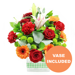 This exuberant mixed bouquet glows with cheerful colour, set against a clean white background that lets every detail shine, as though it were photographed in soft natural light drifting over Stanmore Hill. The arrangement is gathered in a modern, rectangular clear glass vase, the base wrapped in a light green and white gingham ribbon that lends a relaxed, homely charm. At the centre, deep red roses form the emotional core of the design, their tightly layered, velvety petals drawing the eye and conveying classic romance. Encircling them, bright orange gerbera daisies with deep brown centres open out like bright, optimistic faces, offering a strong contrast to the richer reds. Ruffled red-orange carnations are scattered through the bouquet, their frilly petals and rounded forms adding volume, movement, and a softer textural note. Above this vibrant cluster, several white oriental lilies stretch upward; some flowers are fully open, their trumpet-shaped petals marked with delicate pink veins and dusted stamens, while others remain in elegant, closed buds that promise ongoing interest. Between these larger focal blooms, small spheres of lime green pompom chrysanthemums nestle close to the stems, bringing a crisp, contemporary feel and a fresh jolt of colour that keeps the composition light and lively. The flowers are supported by an array of foliage, including broad, glossy dark green leaves and slender stems of silvery eucalyptus that curve gently outward, echoing the natural greenery of Canons Park. In the bottom right of the image, a circular orange graphic slightly overlaps the vase, with the words "VASE INCLUDED" in bold white capitals, clearly indicating that the stylish container forms part of the gift. The finished bouquet feels like a joyful, well-balanced celebration, ready for same-day delivery around Stanmore to mark birthdays, graduations, or housewarmings.