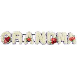 The image shows a tender and elegant floral tribute spelling out GRANDMA in large capital letters, carefully arranged in a straight line against a simple white background so that every detail of the design can be clearly appreciated. Each letter is formed entirely from densely packed white cushion chrysanthemums, the neat, domed flowers arranged so closely that they create a soft, fluffy texture, like a continuous white blanket of petals. Around the edges, a trim of delicate pale lilac ribbon subtly outlines each character, offering a gentle purple sheen that adds refinement and echoes the quiet calm of Stanmore's tree-lined streets. Vivid red highlights are placed thoughtfully throughout the tribute. At the beginning, the letter G is adorned with a compact cluster of deep red roses and a distinctive red-and-white variegated calla lily, its sleek, funnel-shaped bloom curling elegantly and lending a modern twist. The first A features a single, heart-shaped red anthurium lily at its base, the polished surface and central spadix giving a sculptural contrast to the feathery chrysanthemums. In the middle of the word, the letter M is embellished with another group of red roses paired with a second red anthurium, drawing the eye to the centre as a symbol of the grandmother's central place in family life. The final A finishes the sequence with one more red anthurium bloom, creating balance from start to finish. The letters sit on a discreet dark green structural stand that lifts them slightly, providing support without distracting from the flowers. Crafted with care, this personalised funeral arrangement would sit beautifully in a Stanmore chapel or cemetery, offering a heartfelt farewell infused with quiet dignity and enduring love.