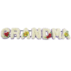 In this image, the word GRANDMA appears as a full-length floral display, set against a pure white background as though prepared for a funeral or memorial service in Stanmore. The seven uppercase letters are meticulously sculpted from masses of white chrysanthemums, each bloom small and tightly packed so that the surface of the letters looks smooth and plush, like a soft bed of petals. A delicate edging of pale lavender or light purple flowers follows the shape of every letter, adding a muted halo of colour that makes the word stand out clearly without breaking the calm, soothing mood. On the first letter, G, three deep red roses cluster toward the lower left curve, their rich, velvety petals and glossy dark leaves providing a warm, heartfelt contrast to the snowy white background. The first A is embellished with two yellow-green cymbidium orchids lying gently along the crossbar; their slightly arching petals and finely speckled throats introduce an air of elegance and individuality. The central R, N, and D are created solely from the white chrysanthemums edged in lavender, keeping the focus on the simple strength of the word itself. Near the right-hand side, the M echoes the beginning of the tribute with another trio of red roses placed in its lower right section, visually linking both ends of the arrangement. The final A repeats the accent of the first A, with two more chartreuse orchids resting among the white blooms, completing the balance of colour and form. All of the letters sit securely on a narrow dark green or black metal frame, which is largely hidden from view but ensures the tribute can be safely displayed at Stanmore churches, cemeteries, and crematorium chapels. The overall impression is one of quiet dignity and deep affection, capturing how a family might honour the guiding presence of a grandmother.