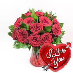 In this romantic arrangement, a generous cluster of deep crimson red roses is presented in a ruby-tinted, fluted glass vase, creating a striking focal point against a clean white background. The bouquet consists of roughly a dozen or slightly more roses, each bloom rich with layered, velvety petals that curl gently away from the centre. Some flowers are fully open, showcasing their intricate spirals, while others remain partially closed, hinting at unfolding emotion-much like a love story growing over time in the quiet, tree-lined avenues of Stanmore. Lush green foliage peeks through and around the roses, providing a fresh contrast; the leaves are smooth, vivid, and slightly glossy, their shapes helping to give the bouquet a natural, rounded silhouette. The vase's ruby-red tone and vertical fluting add sophistication and depth, allowing light to glimmer over its surface in subtle reflections. Through the glass, you can faintly see the stems, criss-crossing in the water, giving a sense of the careful hand-tied structure beneath the artistry. In the lower right foreground, a metallic red heart-shaped foil balloon overlaps the arrangement, bringing a playful, modern twist. Its surface is highly reflective, and in the centre, graceful white cursive lettering spells out I Love You, immediately conveying the sentiment behind the gift. A sleek, silver Cupid's arrow appears to pierce the heart diagonally from lower left to upper right, reinforcing the theme of love and devotion. The whole composition feels tailor-made for anniversaries, Valentine's surprises in Stanmore homes, or any moment when you want to send a clear and heartfelt declaration of affection through flowers.