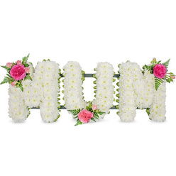 The image portrays a heartfelt floral tribute spelling MOM, carefully photographed against an uncluttered white background so that every detail stands out. Three large capital letters sit side by side, each one built on a sturdy frame and completely blanketed in white chrysanthemums. These are small, spray-type chrysanthemums, packed tightly together so their rounded blooms merge into a soft, even surface that resembles a cushion or a snowy hillside. The pure white petals convey innocence, remembrance, and peace, lending the tribute a quiet dignity that suits a solemn farewell. Along the lower edge of the letters, a slim band of dark green-likely a ribbon over a metal frame-adds a subtle outline and hints at the solid construction beneath the sea of petals. Breaking the expanse of white, three complementary clusters of pink flowers and greenery introduce gentle colour. At the top left of the first M, a standout fuchsia rose sits at the centre, surrounded by paler pink spray roses and rosebuds, with delicate fern fronds adding a lacy, feathered texture. A similar grouping ornaments the lower area between the O and the second M, while another blooms at the top right of the final M, balancing the composition visually. The interplay of bright white and vivid pink creates a sense of warmth within the stillness, evoking gratitude and enduring affection for a beloved mother. This kind of MOM tribute would feel at home in a Stanmore chapel, on a hearse travelling down Marsh Lane, or placed lovingly by a graveside, a soft-spoken yet unmistakable declaration of love expressed through flowers.