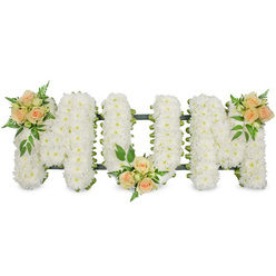 The photograph shows a lovingly designed sympathy arrangement spelling MOM in large, upright block letters, set against a crisp white background that keeps all focus on the flowers. Each letter is completely covered with pure white flowers, almost certainly small spray chrysanthemums, arranged so tightly that they form a smooth, continuous surface with no gaps. The countless little petals create a soft, velvety texture that appears calm and comforting, ideal for a funeral service or celebration of life around Stanmore and the surrounding areas. A neat dark green framework and ribbon edge subtly outline each letter, giving the word strength and clarity while hinting at the skilled construction beneath. Adding warmth and personality, three graceful clusters of spray roses in soft peach and apricot tones are placed along the tribute. One cluster rests on the upper left section of the first M, another nestles at the lower central point of the O, and the third decorates the upper right of the final M, creating a gentle rhythm of colour from left to right. The roses are a mix of buds and partially open blooms, their curved petals catching the light, and each grouping is surrounded by feathery ferns and glossy green leaves for contrast and natural texture. The piece feels both elegant and heartfelt, reflecting the love of children and family members wishing to honour a mother's life in Stanmore, perhaps after a service near the high street or close to Bentley Priory nature reserve. The contrast between the serene white base and the warm peach accents symbolises both peace and enduring affection, making this MOM letter tribute a quietly powerful expression of farewell and remembrance.