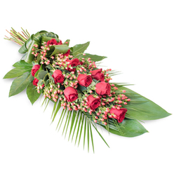 A long, graceful sympathy spray of red roses and berries rests diagonally on a pristine white backdrop, its tapered shape and flowing greenery evoking a calm, contemplative mood. The design is elongated and slightly crescent-like, fuller through the middle and softening as it reaches the lower tip, rather like the gentle curve of the streets around Stanmore Country Park. Along its length, deep red roses form a continuous ribbon of colour, with buds, half-open blooms, and fully unfurled flowers mingling together. The open roses reveal dense, velvety petals layered tightly at the centre, gradually relaxing into softer, looser edges that hint at movement and warmth even in stillness. Interspersed among the roses are generous clusters of pinkish-red Hypericum berries, each berry small, round, and glossy, adding delicate texture and lightness against the saturated crimson tones. Beneath this, a bed of dark green foliage anchors the arrangement: broad, smooth, shiny leaves overlap in a strong, supportive base, while fine, feathery palm fronds reach outward from the lower right side, stretching the silhouette and introducing a flowing, almost draped quality. Subtle accent stems and narrow leaves rise here and there above the main plane, creating a gentle sense of depth without disturbing the calm profile of the spray. At the top left, all stems are neatly aligned and wrapped with a wide band of green leaf, lending a tidy, tailored finish, as you would expect from Flowers Stanmore for a carefully prepared funeral tribute. Lit evenly against the white background, the colours appear true and rich, making this arrangement a meaningful choice for expressing love, respect, and remembrance at funeral services or wakes in Stanmore and the surrounding area.