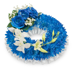 A dignified circular funeral wreath, arranged with a florist's calm precision and photographed on a crisp white background to show every texture and hue. The foundation is a dense carpet of vivid, deep-blue chrysanthemums whose tight, filament-like petals create a richly textured, voluminous ring; the tones move from bright sapphire to midnight blue beneath soft, even lighting. A ruffled white satin ribbon lines the inner rim, offering a satin sheen that offsets the matte, tactile surface of the blooms. Prominently placed at the upper-left quadrant are three large, fully opened blue roses - luxurious and velvety, their layered petals curling inwards like small, serene storms - accented by sprays of delicate white gypsophila and darker green foliage for contrast. Sweeping from the mid-left and curving toward the lower-right, a cascade of ivory white orchids drifts across the wreath, their smooth, almost translucent petals lending an ethereal, quiet elegance. Interspersed among the orchids are fresh green buds, some just beginning to show, suggesting renewal within a tribute. As a local Stanmore florist, Flowers Stanmore often crafts this palette for memorials at nearby churches or services by Stanmore Broadway and Bentley Priory; the colours communicate peace, respect, and enduring memory. The arrangement is tactile and composed - a gentle, emotive statement of condolence suited for funerals, wakes, or commemorative displays where a calm, refined presence is desired.