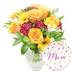 A warm, cheerful bouquet paired with a playful message balloon, arranged and lit to feel like early-afternoon sunshine in a family kitchen. Set against pristine white, the clear glass vase reveals neatly trimmed green stems standing in fresh water, evidence of a recently made arrangement destined for someone special in Stanmore. Dominant peach-orange roses, full and gently unfurling, draw the eye with their soft, petal-rich centres; they give the bouquet a classic, affectionate tone. Interspersed are radiant yellow gerbera daisies whose flat faces and darker centres punctuate the composition with bright optimism. Rich, deep-red or burgundy carnations add a plush, textured counterpoint - their ruffled edges creating shadows and tactile interest - while narrow, speckled white alstroemeria provide small, delicate highlights that lift the palette. The foliage is abundant and varied, from glossy broad leaves that anchor the design to fine, feathery sprigs that add movement and breathe between blooms. Nestled at the base and overlapping the vase, a round white foil balloon carries a scattering of gold and hot-pink polka dots and tiny hearts; across its centre, a lively hot-pink script proclaims best Mum, a personal touch that turns the arrangement into a gift-ready statement. This composition feels particularly suited to deliveries across Stanmore and nearby Canons Park or to a thoughtful surprise left on a doorstep along Stanmore Hill - fresh, heartening, and unmistakably celebratory.