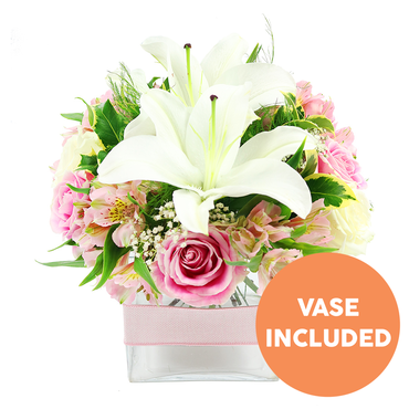 A modern square glass vase brimming with white and pink flowers is shown against a pristine white backdrop, highlighting the clean lines of the design and the softness of the blooms. Dominating the centre are two fully open white Oriental lilies, their broad, smooth petals fanning outward in a gentle curve, each lily accented by prominent deep reddish-brown stamens that lend drama and contrast. Encircling these focal flowers is a generous selection of pink roses, from pale blush tones to a more medium pink, arranged in varying stages of bloom-some tightly furled, others generously open with layered, velvety petals that evoke classic romance. Threaded between the roses are graceful stems of pink and white alstroemeria, their slender petals patterned with fine yellow markings and delicate speckles, contributing detail and a sense of movement. Wispy clusters of white baby's breath fill in the spaces, creating a gossamer, cloud-like effect that lightens the overall look. Around the edge of the arrangement, rich green foliage in multiple shapes-from broad, glossy leaves to lighter, feathery textures-adds contrast and a natural, garden-fresh character, as if the bouquet had been gathered near Bentley Priory Nature Reserve on a spring morning. The stems sit neatly in a clear, cube-shaped vase, with the water visible and the form of each stem adding to the visual rhythm. A pale pink satin ribbon is wrapped smoothly around the vase's middle, offering a soft, decorative touch. In the bottom right corner of the image, a vivid orange circular emblem bears the words VASE INCLUDED in bold white capital letters, making it clear that the vase forms part of the arrangement. The overall impression is of a calm, polished bouquet, ideal for bringing gentle joy to homes and offices across Stanmore.