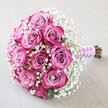 Positioned serenely on a softly textured, cream-toned surface that hints at Stanmore's inviting homes, this lush, hand-crafted bouquet bursts with a sense of romance and celebration. The starring roses, numbering between ten and twelve, display a radiant gradation-from gentle lavender-pink at their heart to vibrant magenta streaks around the edges-unfurling their plush, velvety petals with graceful poise. Nestled among the roses, clouds of fine white baby's breath float delicately, imparting a dreamy airiness that softens the bouquet's rounded form and creates a sense of fullness and movement, as though stirred by a gentle breeze near Stanmore Hill. The stems are tightly gathered and wrapped in shimmering pink satin ribbon, their structure further adorned with a line of dainty white pearls spiraling towards the base to evoke a touch of evening elegance. Peeking from below the ribbon, crisp green and earthy brown stems signal freshness and the artistry of local florists. The arrangement is softly lit, allowing every petal's texture and every Gypsophila floret's detail to shine. Infused with an aura of heartfelt cheer, this bouquet is perfect for a Stanmore wedding, special occasion, or for conveying affection through same-day bouquet delivery in the local community-each element lovingly chosen to capture joy, beauty, and the enduring spirit of Stanmore.