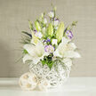 In this image, a carefully composed bouquet of white lilies and lisianthus rests in a sculptural white vase, exuding the understated grace that suits a Stanmore wedding or an elegant home near Canons Drive. At the heart of the display, several lilies are in full bloom, their velvety white petals gracefully arching backwards to reveal speckled centers and bright yellow-orange stamens, while a few upright green buds wait poised to open. Encircling the lilies, lisianthus blossoms in white and the faintest hint of lavender unfold in ruffled layers, with some petals edged in pale violet, giving the arrangement a romantic, almost hand-painted look. Fine sprays of white filler flowers, similar to gypsophila, float like delicate clouds between the larger blooms, supported by slender strands of dark green foliage and touches of deep blue-purple flowers that tuck into the arrangement, adding pockets of rich colour. The flowers rise from a rounded vase fashioned from interlocking white strands, creating an open, basket-like pattern that feels light and modern, much like the bright, airy living rooms overlooking Stanmore Country Park. On the smooth white surface beside the vase, two decorative woven spheres in natural beige tones echo the same intricate, rattan-style texture, adding an earthy note without disturbing the arrangement's serenity. Behind, a softly lit beige background with a fine vertical weave acts as a neutral canvas, creating gentle shadows and highlighting the crisp outlines of each petal. The overall impression is one of calm, polished beauty, perfect for sending heartfelt condolences, marking an anniversary, or arranging same-day delivery across Stanmore from Flowers Stanmore when you want flowers to speak with quiet clarity.
