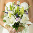 For a wedding morning framed by pale light, this hand-tied bouquet presents a study in graceful contrasts: large, star-shaped lilies in creamy white form the composition's focal chorus, their opened faces polished and softly luminous. Around them, lisianthus edged in dusky violet introduce a cool, romantic counterpoint; their ruffled petals and gently curled buds add depth and a painterly texture that feels both timeless and contemporary. Sprays of tiny baby's breath float between the main blooms like delicate mist, giving the arrangement an ethereal lightness, while slender green threads - almost calligraphic in their movement - outline and lift the bouquet so it reads airy from every angle. Under gentle daylight the flowers glow with a soft halo; highlights on the lilies' petals and the subtle purple rims of the lisianthus create quiet focal points that guide the eye through the design. There is an understated aroma: the creamy sweetness of lilies tempered by fresh, grassy notes from the foliage, a scent I associate with early ceremonies near Canons Park or the quieter streets off Marsh Lane. As a local florist I arrange each stem with purpose so every element is felt - the lilies' structural grace, the lisianthus's romantic edge, the baby's-breath's innocence - balancing form and movement. Flowers Stanmore often crafts bouquets like this for couples celebrating at nearby venues; the effect is refined, intimate, and designed to be both seen and remembered, a floral promise carried close to the heart on a day of new beginnings.