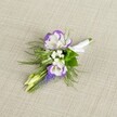 A delicately crafted floral boutonniere lies against a softly textured, light beige linen-style background, the neutral fabric allowing every petal and leaf to stand out with quiet clarity. At the heart of the design sit two white lisianthus blooms, their petals gracefully ruffled and edged with a rich, velvety purple that deepens toward the tips, as if hand-painted. Nestled close to these focal flowers are fine clusters of tiny white filler blooms, reminiscent of baby's breath, adding a cloud-like softness and gentle volume. Running vertically through the composition is a slender spike of deep purple grape hyacinth, or muscari, its tightly packed, bell-shaped flowers adding a distinctive, textural accent and echoing the purple tones of the lisianthus. Around the flowers, airy, feathery greenery-much like asparagus fern-fans out in light, lacy fronds, joined by slightly broader, deeper green leaves that anchor the piece and lend a natural sense of depth. Two pale green buds remain partially closed, hinting at freshness and new beginnings, like a spring morning walk through Stanmore Country Park. The stems are neatly gathered and wrapped with a crisp white satin ribbon, smoothly wound and finished in a small, elegant bow, clearly prepared to be pinned to a lapel. The mood of the image feels serene and sophisticated, ideal for a groom or groomsman heading to a Stanmore wedding near Canons Park or a formal celebration in one of the area's historic halls, where the understated colours and careful craftsmanship quietly highlight the significance of the day.