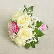 A small, hand-tied bouquet is laid diagonally across a neutral, light beige fabric, whose fine, almost linen-like weave gives a natural, slightly rustic texture beneath the flowers. The focal point is a cluster of five creamy white or ivory roses, each bloom open enough to display layered, spiralling petals that feel plush and soft to the eye. Nestled near the front-left of the posy is a single, vivid fuchsia-pink rose, its richer colour immediately drawing attention and adding a lively spark to the otherwise gentle palette. Threaded between every rose are airy sprays of white baby's breath, their tiny blooms forming delicate clouds that create a sense of lightness and movement, as though a soft Stanmore breeze had just brushed over the flowers. Around the outer edges, sleek, deep green leaves-likely ruscus, commonly used in local arrangements for its durability-provide contrast and definition, framing the roses and baby's breath with a natural, verdant border. The stems are neatly aligned and wrapped in a bright pink satin ribbon, its silky surface smooth and reflective, forming a polished handle. Over this ribbon, multiple narrow rows of small, iridescent white pearls have been meticulously placed, suggesting the care of an experienced florist who might be preparing this for a wedding near Stanmore Country Park or a prom at a local school. Soft, even lighting washes gently over the bouquet, revealing every nuance of cream, pink, and green, while the overall composition radiates quiet celebration, tenderness, and an intimate sense of occasion suitable for corsages, boutonnieres, or petite centrepieces.