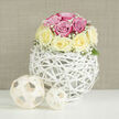 This image captures a serene still life of a contemporary floral design, carefully composed on a clean, bright white tabletop that reflects the gentle light. Dominating the centre is a dome-shaped bouquet of roses, arranged so precisely that the blooms form an almost perfect sphere. The rose petals blend shades of soft, romantic pink with creamy pale yellow, each blossom opening generously, as if caught just after being delivered fresh from a local Stanmore florist. Between the larger heads of the roses, delicate sprays of white baby's breath weave through the arrangement, their countless miniature blossoms creating a cloud-like border that softens the composition and introduces a hint of countryside charm, reminiscent of a peaceful walk through Bentley Priory Nature Reserve. Subtle green foliage peeks out at the base, providing a natural transition into the vessel. The flowers are nestled into a striking spherical vase fashioned from thin, white rattan-style strands that twist and cross over one another, forming an open, basket-like structure. This airy design contrasts with the fullness of the blooms, adding a sense of lightness and modern artistry, like a sculptural piece in a contemporary Stanmore home. On the same surface, close to the vase, sit two smaller decorative spheres made from natural, light beige wooden strips, their woven patterns echoing the texture of the main container. The background is a calm, neutral beige with a fine horizontal texture that resembles linen, giving the entire scene a warm yet minimalist feel. The image suggests a refined centerpiece for intimate gatherings, engagement celebrations, or thoughtful thank-you gifts, beautifully suited to same-day delivery across Stanmore and the surrounding North West London neighbourhoods.