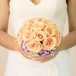 A close-up, chest-level photograph shows a bride standing calmly, her torso framed against a soft, pale background that feels like gentle daylight over Stanmore. She wears a classic white wedding gown with an ivory undertone, the bodice delicately traced with lace and a modest V-neckline that adds quiet elegance. The fabric of the skirt falls in soft folds beneath her hands, creating a subtle, flowing texture that contrasts with the structured beauty of the bouquet she holds. Cradled carefully between her lightly manicured hands is a perfectly round bridal bouquet, its focal point a dense cluster of roses in warm peach, light apricot, and blush pink. The petals are tightly layered, forming soft spirals that suggest both freshness and intimacy, as though the roses were gathered that morning from a local Stanmore garden. Encircling the base of the peach roses is a striking ring of ruffled blooms, likely lisianthus, their petals deep purple at the edges fading gracefully into creamy white centers. This vivid border adds depth and drama without overpowering the gentle romance of the roses. Wisps of tiny white filler flowers, reminiscent of baby's breath, are tucked between the purple edges and the underside of the bouquet, creating a cloud-like softness and a classic bridal touch. The bride's nails are finished in a pale, neutral shade with a hint of glitter on one thumb, suggesting a personal, modern twist. Together, the gown and bouquet evoke a sense of poised anticipation, ideal for a Stanmore wedding ceremony, and beautifully capture the timeless, handcrafted style that local florists bring to intimate celebrations across the area.
