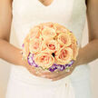 This bridal bouquet reads like a soft poem in peach and violet: a symmetrical cluster of apricot roses at the centre, each bloom tightly cupped and glowing with creamy warmth, surrounded by a circlet of purple-tinted lisianthus that offers a cool, refined counterpoint. The lisianthus frames the roses like a delicate collar, its white inner petals lifting the apricot tones and lending an ethereal calm. Interwoven between the main blooms, a fine lacework of tiny white filler flowers creates a whisper-light texture, making the whole arrangement feel buoyant and almost weightless when held. The portrait lighting is gentle and even, bathing petals in a warm glow that highlights the veins and the subtle gradations of colour from centre to edge. Texturally, velvet and whisper meet: the roses bring creamy plushness, the lisianthus a silky crispness, and the filler's tiny stars add a slight, breathy crispness at the margins. The bouquet's emotional language is classic and hopeful - perfect for a ceremony near the green lanes of Stanmore Broadway or a small reception at a local hall in Harrow Weald. As an experienced florist describing this piece, I notice the careful rhythm of placement and the reassuring weight in the stems that helps the bouquet sit naturally in a bride's hands. It's thoughtful, refined, and designed to read beautifully in close-up photos and quiet memories alike, a gentle centrepiece for a day of promises and tender celebration.