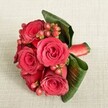 A richly romantic bouquet of deep red roses and berries rests on a soft, light beige textured fabric, captured from a slightly elevated angle that shows every velvety petal in detail. Around five or six lush roses, each in full bloom, form the heart of the design, their petals spiralling from tight, shadowed centres to open, gently ruffled edges that glow in shades of crimson and scarlet. Threaded between the blooms are clusters of small, rounded Hypericum-style berries in tones of ruby red and reddish-pink, their smooth, bead-like surfaces adding a playful contrast to the softness of the roses. Encircling the flowers, broad, dark green tropical leaves are artfully curved and folded, creating a polished, collar-like frame that holds the composition together and highlights the rich colours at the centre. Near the base, a band of matching red satin ribbon wraps the stems with a subtle sheen, suggesting expert hand-tying and a finish suitable for a wedding, prom, or special celebration in Stanmore. The neutral cream and beige background, with its fine woven texture, gently diffuses the light and makes the reds and greens appear even more vivid, as if the bouquet has just been set down on a linen-covered table before being pinned as a corsage or worn as a statement boutonnière at a local ceremony. The overall impression is one of intimate luxury and heartfelt romance, the kind of detailed floral work a Stanmore florist would create for a couple celebrating an anniversary dinner or a formal event near Bentley Priory or along The Broadway, where every petal and berry has been placed with care and intention.