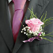 This image shows a single pink rose boutonniere pinned carefully to a charcoal-grey suit lapel, the kind of refined detail often seen at weddings and formal celebrations across Stanmore. The rose itself is at that perfect stage of bloom-open enough to reveal its spiral of petals, yet still compact, with edges that curl softly rather than splay. Its colour is a warm, gentle pink that suggests tenderness and understated romance, not unlike the soft evening sky over Stanmore Hill. Around the rose, feathery green foliage extends in slender arcs, their fine, needle-like leaves introducing a light, natural texture that contrasts with the crisp cut of the suit. At the base and nestled between the greens, clusters of white gypsophila gather like tiny snowflakes, the multitudinous small blossoms forming an airy, cloudlike border that highlights the rose's blush tones. The stems are wrapped tightly in lilac satin ribbon, smoothed and tied by skilled hands so that every fold lies flat and even, catching faint reflections in the ambient light. The photograph feels intimate, as though captured while someone adjusts their jacket before stepping out onto Marsh Lane or heading toward a local Stanmore synagogue or church for the ceremony. The juxtaposition of textures-firm fabric, soft petals, crisp baby's breath, and sleek ribbon-creates a tactile richness you can imagine even without seeing. The boutonniere conveys quiet pride and a sense of occasion, a small floral emblem that marks the wearer as part of something joyful and significant, thoughtfully crafted and placed close to the heart.