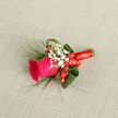 This boutonniere is a studied balance of colour and texture, centred on a fierce magenta rose whose petals appear velvety and full of life, opening to reveal that intimate spiral at the heart. Sweeping loops of deep, glossy foliage - reminiscent of folded aspidistra - form a smooth, structured backdrop, while wispy grassy sprigs breathe an effortless, windswept quality into the design. Tiny clusters of white gypsophila sit like soft stars around the bloom, bringing a purity and old-fashioned romance, while several plump, red hypericum berries reflect the rose's tone and add a festive, tactile gloss. The stems are professionally bound with a bright red satin ribbon, wound snugly and finished with a neat bow that both secures the work and reads as a deliberate stylistic choice. Laid against a light beige, linen-like fabric, the arrangement's colours sing with refined contrast and depth. As a local florist in Stanmore, I picture this boutonniere walking down the high street near Stanmore Broadway or being chosen for intimate ceremonies at local chapels and community halls, and Flowers Stanmore can prepare similar pieces for same-day delivery across Stanmore and the Harrow area. The overall mood is joyous and assured - an elegant little bloom that feels handcrafted, wearable and perfect for milestones where warmth and quiet sophistication are desired.