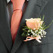 The charm of this boutonniere is in its gentle balance and thoughtful craftsmanship, a single peach rose held like a small sun against a dark suit lapel. Its petals move from creamy blush at the edges to a warmer coral heart, each layer catching light in a way that makes the bloom appear almost luminous. Surrounding the rose, delicate sprays of baby's breath float like tiny clouds, their white points softening the composition and lending an ethereal texture. Wisps of fresh green foliage-slender, slightly serrated leaves and a curled support leaf-create structure and a hint of wildness without overpowering the central bloom. A satin ribbon, matched to the tie's coral-pink sheen, is tied with a precise little bow at the base, a florist's signature of care that keeps the arrangement neat and wearable. Against charcoal tailoring, the colours pop with a refined warmth; the ensemble reads as both celebratory and quietly sophisticated. Close to home in Stanmore and Harrow, where morning light filters through trees on Marsh Lane and local couples stroll past The Broadway, this boutonniere would be a fitting, fragrant finishing touch. At Flowers Stanmore we imagine it pinned on a groom or cherished guest-the scent, the texture, and the timeless palette all working together to mark a significant, joy-filled day.