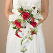 In this photograph, the camera lingers on a sophisticated cascading bridal bouquet held securely at the bride's waist, the upper part of her white gown framing the flowers with gentle curves of fabric and lace. The design is a striking composition of red and white, beginning with a core of deep crimson roses, each bloom richly layered and fully opened, conveying passion and enduring love. Nestled among them are elegant white lilies, their wide, porcelain-like petals curling slightly at the edges and revealing fresh green stamens that hint at a light, classic fragrance. From the central cluster, white Phalaenopsis orchids sweep downward, creating a soft waterfall that defines the bouquet's silhouette; their silky petals and small buds form graceful lines that echo the fall of the bride's dress. Between the main blooms, delicate sprays of tiny white baby's breath float like a fine mist, softening the strong colour contrasts, while bright red berries - likely hypericum - pop up throughout the greenery, adding cheerful bursts of texture and seasonal charm. A variety of foliage, from slender fern fronds to broader, glossy leaves and tiny green-tipped white florets, surrounds and supports the flowers, giving the bouquet depth, movement, and a natural, garden-inspired feel reminiscent of Stanmore's tree-lined avenues and open green spaces. The bride's gown shows subtle embroidered or lace detailing that mirrors the orchids' refinement. Behind her, a blurred backdrop of warm beige tones ensures that the eye stays firmly on the bouquet. The overall effect is one of classic romance with a luxurious twist, ideal for weddings across Stanmore, from historic churches to modern local venues.