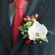 This refined buttonhole is described as if by a local Stanmore florist who has threaded ribbons for wedding mornings on Watling Avenue and deliveries to Canons Park: a single white Phalaenopsis orchid claims the centre, its broad, luminous petals soft as velvet to the eye and set off by a labellum streaked with yellow-gold and cream. The orchid's smooth, sculptural form is grounded by glossy, deep green foliage that forms a low, supportive collar, while tiny clusters of gypsophila drift through like a pale, celebratory haze. Plump hypericum berries-warm coral with a delicate blush-provide rounded punctuation, catching highlights that seem to mirror the sheen of a solid red satin tie nearby. The suit is a dark charcoal jacket with a prominent left lapel; beneath it, a pale blue-grey patterned shirt peeks through, offering quiet contrast to the tie's confident crimson. A slender red ribbon is looped and tied at the boutonnière's base, tidy and intentional, harmonising with the tie and adding a final, crafted flourish. The image reads as a moment of preparation: cool, late-morning light, the mild scent of orchid and leaf, and the anticipation of vows or a joyous reception. Flowers Stanmore would describe this piece as ideal for grooms, fathers of the bride, or anyone marking a milestone in the local area-ready to be pinned before photos at the church on Stanmore High Street or a reception near Canons Park, made to look effortless and kept fresh until the first applause.