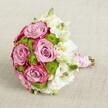 A hand-tied bridal bouquet rests on a softly textured, light beige fabric, its rounded silhouette captured from a gently elevated angle that shows off every curve and petal. At the centre, large, fully opened roses in a soft lavender-pink unfurl like satin, their inner petals pale and romantic, deepening towards the outer edges into a richer, almost fuchsia tone. Between these glowing pink roses, clusters of bright chartreuse button chrysanthemums nestle closely, their tiny, tightly packed petals creating a playful, almost velvety texture and a lively pop of green that wakes up the whole design. Threaded throughout are delicate white blooms, most likely lisianthus, with layered, ruffled petals and soft greenish-yellow centres that lend a classic, almost vintage elegance. A few closed buds peek through, signalling the bouquet's freshness, as if it's just been finished in a Stanmore florist's studio that morning. The stems are neatly gathered and bound into a slim handle, wrapped carefully with smooth, pink satin ribbon. Over this, a fine strand of small white pearls spirals gracefully, like heirloom jewellery, catching the light in subtle highlights. The overall mood is romantic yet polished, perfect for a bride walking down the aisle at a ceremony near Stanmore Country Park, or for an intimate celebration at a local restaurant. The mix of modern chartreuse green, timeless pink roses, and soft white accents, set against the neutral background, creates a bouquet that feels both contemporary and deeply sentimental, ideal for wedding inspiration or elegant gift-giving across Stanmore and the surrounding North West London neighbourhoods.