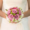 Held close against the intricate lace of a bridal gown, this bouquet captures the warm, heartfelt mood of a Stanmore wedding morning. Lush roses in a shade that drifts between lavender-pink and muted mauve take centre stage, their petals unfolding in velvet-soft layers that create depth and movement. Between them nestle lime-green button chrysanthemums, each bloom a tidy, rounded pompom that adds a spirited burst of colour and a crisp, contemporary feel. The playful green pairs beautifully with the more romantic roses, making the bouquet feel both joyful and poised. Along the outer edge, creamy white blossoms and pointed ivory buds form a light, cloud-like halo that softens the palette and ensures every shade can sing without clashing. The bouquet is tightly hand-tied, resting comfortably in the bride's hands with just enough weight to feel reassuring as she walks the aisle of a local Stanmore church or civil venue. You can almost sense the fragrance rising up: the familiar perfume of roses mingled with fresh, green notes from the chrysanthemums and foliage, something calm and uplifting that follows the bride from photos in Canons Park through to the reception. Gentle daylight caresses each flower, throwing subtle shadows and highlights that draw the eye from one petal to the next. Created with an experienced florist's understanding of balance and longevity, the bouquet is built to remain radiant throughout the day, from the first look to the last toast, quietly expressing joy, hope, and the beauty of new beginnings.