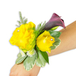 A meticulously arranged wrist corsage shown diagonally across a light-to-medium forearm against a crisp white background, radiating cheerful elegance for prom, wedding or formal wear. The focal pair of roses are a buttery, golden yellow - one fully open and softly ruffled on the left, the other a slightly smaller, cupped bud to its right - their velvety petals suggesting a gentle sweet-rose scent. Each rose is finished with a single clear rhinestone nestled near its heart, catching studio light and sending tiny pinpricks of sparkle across the petals. Behind and rising just above the roses, a single slender calla lily unfurls in a deep plum-to-burgundy tone, its smooth trumpet shape and satiny surface providing a dramatic, sophisticated counterpoint to the sunny roses. Surrounding these blooms, feathery asparagus fern creates airy movement and soft texture, while broader variegated leaves with pale, creamy edges (reminiscent of pittosporum) form a stable, glossy base. Little clusters of white button-like filler flowers - suggestive of spray chrysanthemums or tiny baby's breath - tuck into the composition to add fullness and delicate contrast. The corsage is fastened with a slim, mostly concealed band so the floral composition sits comfortably on the wrist, angled from lower left of the frame toward the centre right, clearly demonstrating how it would be worn. The clean white backdrop makes colour and detail read clearly for anyone relying on tactile or verbal guidance; the overall impression is joyful yet refined, a wearable accent that pairs sunshine-yellow optimism with evening-plum sophistication - a design we often bring to bridesmaids and prom-goers from the Stanmore area for memorable celebrations.