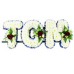 In this image, the name TOM is presented as a refined floral sympathy tribute, formed from a trio of substantial block letters that rest in a neat line. Each letter is densely filled with white chrysanthemums, their petals overlapping to create a soft, plush surface that feels gentle and comforting, much like the atmosphere of a thoughtfully arranged Stanmore funeral service. The outer edges of the letters are framed with a royal blue satin ribbon, pleated and fixed with precision so that it catches the light and adds a stately outline to the pure white blooms. At selected points on the T, O, and M, small yet striking groups of accent flowers interrupt the white expanse: velvety deep red roses, refined cream and white spray roses, and slender white flowers with a freesia-like elegance. These are surrounded by rich green foliage and feathery ferns, creating a natural, garden-like feel that brings warmth and personality to the tribute. The letters are supported by a slim dark bar running beneath them, subtly visible and clearly designed to provide a stable base for display on stands, a hearse, or within a chapel setting in and around Stanmore. The composition of white, blue, and red not only offers pleasing visual balance but also carries a sense of purity, peace, and enduring love for Tom. The tribute feels calm, composed, and deeply respectful, as if it has been created to speak quietly on behalf of family and friends when words are too difficult, offering a final gesture of care through carefully chosen flowers.
