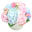 Set against a bright white background, this round white hatbox arrangement radiates soft elegance and abundance. The box is smooth and uncluttered, with a faint ridge around its middle that subtly echoes the rounded shape of the bouquet above. Overflowing from the top is a lush dome of pastel flowers, dominated by large, ruffled pink peonies whose layered petals look almost like folds of satin. Some of the peonies are fully open, displaying their generous centres, while others are still in bud or just beginning to unfold, creating a gentle rhythm of shapes and stages. Nestled among them, powder-blue hydrangea heads provide a wide, cloudlike base, each composed of many tiny blossoms that together give a feeling of lightness and volume. Delicate light pink spray roses are tucked into the gaps, their small, tight buds and semi-open blooms adding intricate detail and the sense of a bouquet that will continue to evolve over several days. Rising above this soft sea of rounded petals, two sleek pink calla lilies add a modern note-one arcs from the upper left of the arrangement, the other from the right-each with a smooth, trumpet-shaped form and a slightly deeper, more vivid pink. Sparse touches of rich green foliage peek out near the base, providing just enough contrast to frame the pastels. The overall effect is one of calm luxury and gentle joy, perfect for a new baby celebration in Stanmore, an intimate birthday lunch near Canons Park, or a thoughtful, soothing delivery to someone in the local community who would be comforted by a cloud of soft blues and pinks arriving at their door.