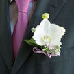 Pinned just above the suit pocket, this boutonniere centres on a radiant white orchid, glowing softly against a charcoal lapel like a point of light on an evening stroll near Stanmore Station. The orchid's petals open in a calm, confident curve, their texture smooth as fine china with the faintest ivory hue tracing the outer edges, adding warmth and depth. Encircling the bloom, tender green leaves and small lilac flowers provide contrast in both colour and feel, mirroring the quiet, leafy streets that wind around Stanmore's residential avenues. A narrow violet ribbon binds the stems in a clean, tailored finish, its sheen gently reflecting the richer lilac shade of the tie below. The result is a composition that looks measured and thoughtful, a small but striking focal point chosen with real care-exactly the kind of detail couples often seek when visiting Flowers Stanmore to discuss their wedding flowers. The dark suit fabric creates a strong, structured stage for the boutonniere, allowing the white orchid to appear almost weightless, as if hovering just above the lapel. There is a palpable sense of anticipation in the image: perhaps this is moments before stepping into a ceremony at a Stanmore synagogue, a local church, or a registry office nearby. Every element appears freshly prepared-the petals firm and hydrated, the lilac blooms vibrant, the ribbon smooth and uncreased-suggesting same-day craftsmanship by a skilled florist. More than an accessory, this boutonniere feels like a small emblem of celebration and affection, quietly sharing in the joy of a significant day in Stanmore.