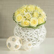 This arrangement offers a delicate study in light and texture: creamy yellow roses gathered into a compact, domed bouquet with ruffled centers that open like soft sighs, their warm tones tempered by a halo of tiny white gypsophila that seems to float among the blooms. The bouquet rests in a sculptural white wicker orb whose airy lattice provides a tactile contrast to the petal softness and lends the presentation an architectural clarity. Nearby, low-profile woven spheres provide a playful, contemporary motif that echoes the basket's material while keeping the overall composition clean and modern. Carefully curled blades of greenery encircle the rim, their gentle loops creating subtle movement and framing the flowers with considered restraint. In the right light-early morning or late afternoon-the petals glow faintly and the mix of rose and gypsophila gives off a scent like sun-warmed linen and freshly cut grass, with a whisper of earth that grounds the sweetness. Suitable for an understated celebration, a thoughtful congratulations, or as a calming centrepiece, this design reflects an experienced florist's eye: balanced proportions, clean lines, and textural contrast. As a Stanmore-based maker I often deliver similar pieces to homes off Stanmore Broadway and to local gatherings near Stanmore tube; it's the kind of bouquet that settles naturally into domestic interiors, adding a touch of quiet elegance and an approachable modernity.