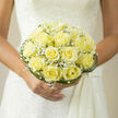 Held gently at the waist of a bride in classic white lace, this bouquet of pale yellow roses reads like a quiet promise: fresh, elegant, and intimately joyful. The roses are a gentle butter-cream hue, each bloom opening into a layered rosette with velvety petals that reflect a soft, natural light-silky to the eye and substantial to the touch. Interspersed through the cluster, pockets of white baby's breath create a frothy halo, their tiny blossoms offering an ethereal, cloud-like contrast that lifts the composition. The bouquet's silhouette is refined and compact, framed by looping strands of glossy green grass that curve around the blooms to give a sculptural border and a contemporary twist. A tidy green collar finishes the base, suggesting garden-fresh crispness beneath the lush flowers. Close your eyes and you can imagine a faint, powdery fragrance where light honeyed notes from the roses mingle with the clean greenness of foliage, a scent both nostalgic and quietly optimistic. As someone who arranges wedding bouquets for ceremonies at venues around Stanmore Tube and the leafy lanes toward Canons Park, I picture this design paired with soft morning light or the warm glow of an intimate evening-an arrangement that whispers of devotion, tenderness, and the simple beauty of a new beginning. Flowers Stanmore crafted with care, it's designed to complement rather than compete with the bride's gown, offering a tactile memory to hold on a day that matters.