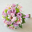 Displayed against a soft, light grey surface with a subtle linen texture, this hand-tied wedding bouquet appears both polished and romantic, as though prepared moments ago by a Stanmore florist for a bride stepping out of a classic townhouse. The arrangement forms a dense, rounded posy, its structure centred on generous lavender roses whose petals curl open in velvety spirals, each layer overlapping the next in a rich, opulent texture. Among the roses, pink alstroemeria blooms weave gracefully, their slender petals washed in blush, cream, and hints of gentle yellow, with delicate darker flecks near their throats that create a sense of movement and detail up close. Clusters of vivid chartreuse button chrysanthemums sit like small, structured pompons, their crisp, green orbs injecting a lively contrast that keeps the palette fresh and contemporary. Scattered through the bouquet are fine sprigs of white baby's breath, the tiny blossoms forming airy halos that soften the boundaries between flowers, adding a dreamy, cloud-like atmosphere. The stems are neatly bound at the base with a broad band of satin ribbon in a warm pink shade, its glossy surface reflecting the light subtly, while a strand of small white pearls is wrapped around the handle, echoing the jewellery often worn for weddings in and around Stanmore. Below, the natural stems extend in a clean, trimmed bundle, showing woody textures and green hints that ground the romantic design. The bouquet feels perfect for walking down the aisle at a ceremony near Stanmore Common or for same-day delivery to mark anniversaries and new beginnings throughout the local area.